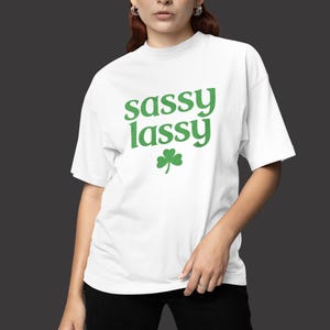 May include: White t-shirt with the green text "sassy lassy" and a green shamrock. The shirt is worn by a person wearing black pants. The background is a dark gray.