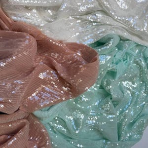 Puede incluir: Primer plano de tela brillante en tonos rosa rubor, blanco y verde menta. La tela está cubierta de pequeñas lentejuelas reflectantes, creando una apariencia texturizada y glamurosa. Los pliegues y drapeados de la tela añaden profundidad e interés visual.