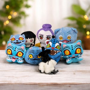 Pode incluir: Uma coleção de brinquedos de pelúcia com vários personagens de desenhos animados. Os brinquedos incluem criaturas azuis semelhantes a gatos com olhos amarelos e dentes afiados, e outras bonecas com designs únicos. Os bichos de pelúcia estão dispostos em uma superfície de madeira clara.