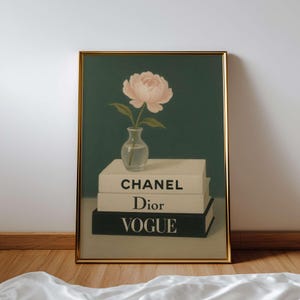 May include: A framed art print featuring a pink peony in a small glass vase atop a stack of books. The books are labeled "CHANEL", "Dior", and "VOGUE". The background is a dark teal color, and the frame is gold.