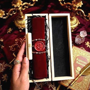 Puede incluir: Una caja de madera con un pergamino de terciopelo rojo, sellado con un sello de cera de rosa y una cadena de perlas. Un pergamino enrollado con un diseño de rosa está al lado del pergamino de terciopelo. La caja está abierta, revelando un interior negro. Se ve un anillo de oro.