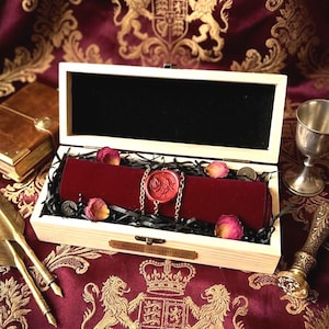May include: A rectangular light-colored wooden box with a hinged lid containing a dark red velvet scroll secured with a gold chain and a red wax seal featuring a rose design. The box has a metal plaque reading BY ROYAL COMMENDATION.
