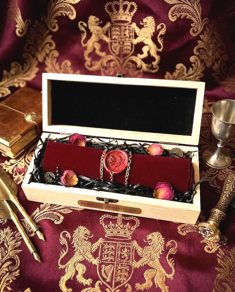 May include: A rectangular light-colored wooden box with a hinged lid containing a dark red velvet scroll secured with a gold chain and a red wax seal featuring a rose design. The box has a metal plaque reading BY ROYAL COMMENDATION.