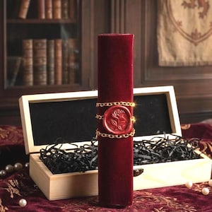 May include: A cylindrical scroll wrapped in deep red velvet, secured with a gold-toned metal chain and a circular red wax seal featuring an embossed rose design. The scroll stands upright in an open, light-colored wooden box filled with black paper.