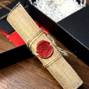 K&ouml;nnte beinhalten: Eine gerollte Pergamentrolle, mit Schnur gebunden und mit einem roten Wachssiegel versehen. Der Stempel tr&auml;gt die Aufschrift "With Love". Die Rolle besteht aus einem stoff&auml;hnlichen Material und wird in einer schwarzen Geschenkbox pr&auml;sentiert.