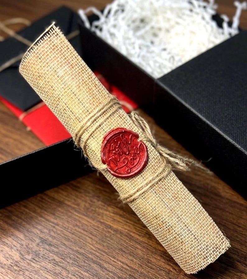 K&ouml;nnte beinhalten: Eine gerollte Pergamentrolle, mit Schnur gebunden und mit einem roten Wachssiegel versehen. Der Stempel tr&auml;gt die Aufschrift "With Love". Die Rolle besteht aus einem stoff&auml;hnlichen Material und wird in einer schwarzen Geschenkbox pr&auml;sentiert.