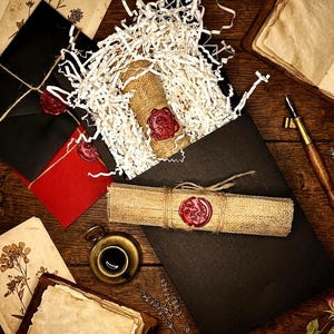K&ouml;nnte beinhalten: Eine rustikale Szene mit einer schwarzen Box, die einen mit Jute umwickelten Gegenstand enth&auml;lt, der mit rotem Wachs versiegelt ist. Eine Schriftrolle, Umschl&auml;ge, eine kleine Tasse und ein Stift sind ebenfalls vorhanden, alles auf einer Holzoberfl&auml;che angeordnet.