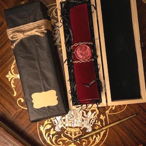 May include: A dark red velvet scroll secured with a gold chain and a red wax seal featuring a rose design, nestled in a wooden box with black filler. Beside it sits a black paper-wrapped package tied with twine and a gold-toned fountain pen.