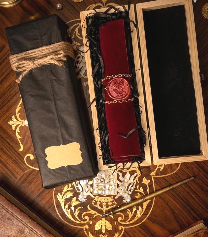 May include: A dark red velvet scroll secured with a gold chain and a red wax seal featuring a rose design, nestled in a wooden box with black filler. Beside it sits a black paper-wrapped package tied with twine and a gold-toned fountain pen.
