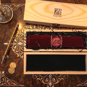 May include: A wooden box containing a dark red velvet scroll secured with a rose-embossed wax seal and a metal chain. The box lid features the text GoldFrame Vintage Note. A brass fountain pen and two gold coins are placed on the table nearby.