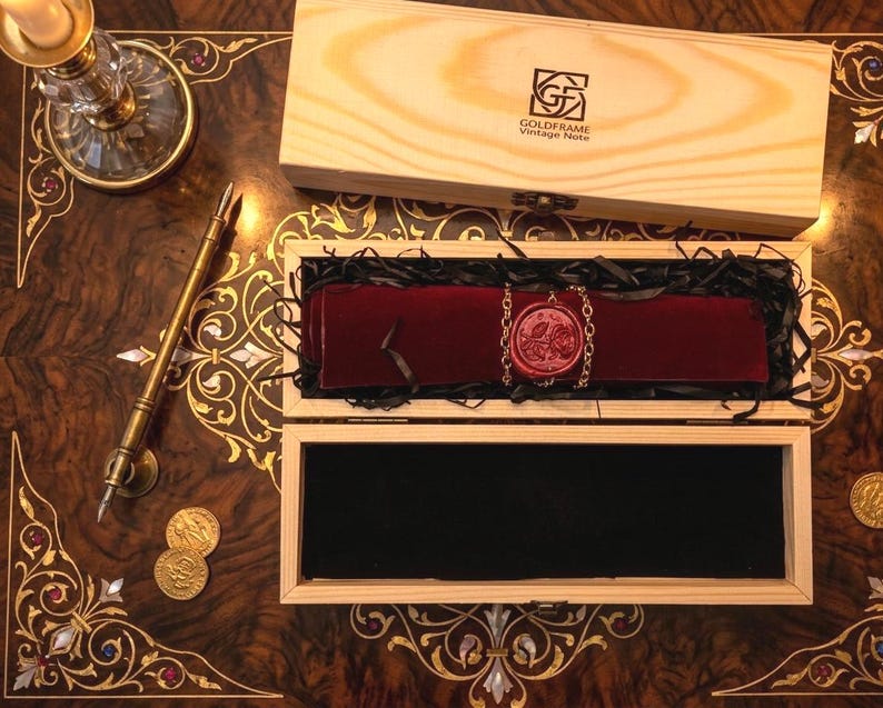 May include: A wooden box containing a dark red velvet scroll secured with a rose-embossed wax seal and a metal chain. The box lid features the text GoldFrame Vintage Note. A brass fountain pen and two gold coins are placed on the table nearby.