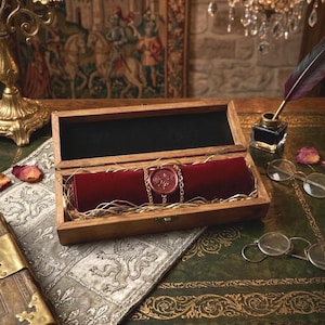 May include: A rectangular wooden box containing a dark red velvet scroll secured with a gold chain and a circular wax seal featuring a rose design. The scroll rests on a bed of straw-like filler inside the open box, which has a dark interior lining.