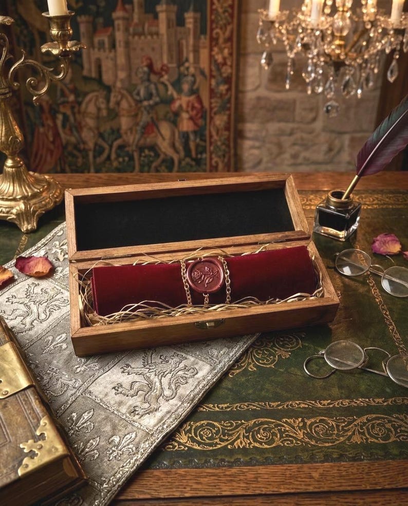 May include: A rectangular wooden box containing a dark red velvet scroll secured with a gold chain and a circular wax seal featuring a rose design. The scroll rests on a bed of straw-like filler inside the open box, which has a dark interior lining.
