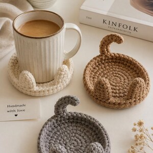 May include: Three circular crochet coasters in cream, tan, and gray, each featuring two small upright ears and a curved tail. One cream coaster supports a ribbed ceramic mug filled with coffee. A small white card reads Handmade with love.