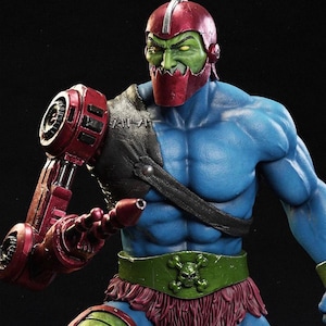 May include: A detailed action figure of a muscular character with a green face, red helmet, and blue body. The figure has a mechanical red arm and a green belt with a skull and crossbones design. The figure is a collectible.