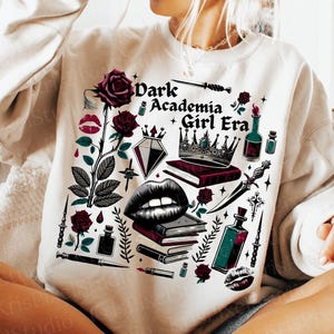K&ouml;nnte beinhalten: Cremefarbenes Sweatshirt mit einem grafischen Design, das den Text "Dark Academia Girl Era" und Illustrationen von Rosen, B&uuml;chern, einer Krone und anderen gotisch inspirierten Elementen zeigt. Das Sweatshirt hat eine lockere Passform.