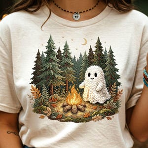 May include: A cream-colored t-shirt featuring a graphic of a friendly ghost sitting by a campfire in a forest. The illustration includes green pine trees, a crescent moon, and autumn leaves. The ghost is white and appears to be knitted.