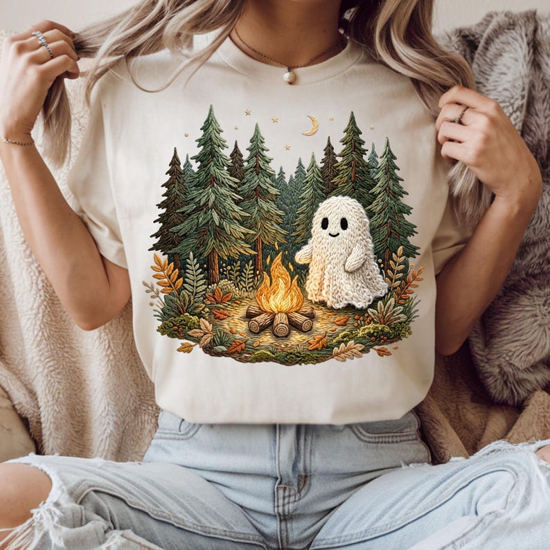 May include: Cream-colored t-shirt featuring a whimsical illustration of a friendly ghost near a campfire in a forest setting. The design includes pine trees, a crescent moon, and autumn foliage, creating a cozy, outdoorsy theme.