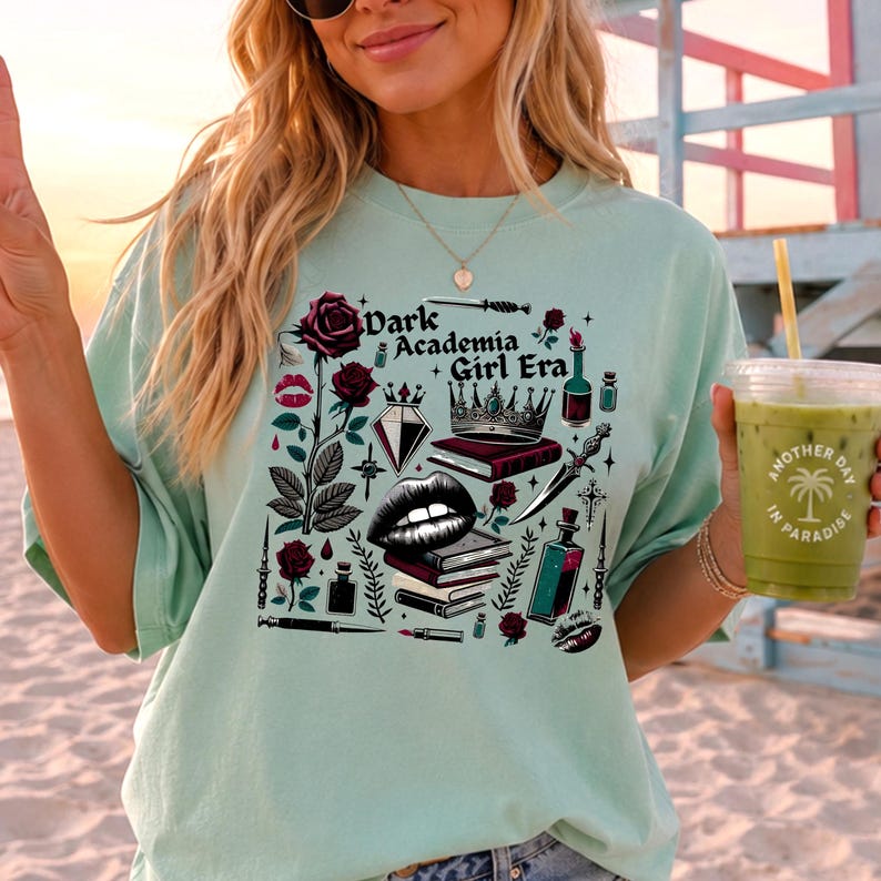 K&ouml;nnte beinhalten: Hellgr&uuml;nes T-Shirt mit einem grafischen Design mit dem Text "Dark Academia Girl Era" und Illustrationen von Rosen, B&uuml;chern, einer Krone und anderen gotisch inspirierten Elementen. Die Person h&auml;lt ein gr&uuml;nes Getr&auml;nk.