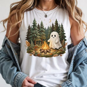 May include: White tank top featuring a whimsical illustration of a friendly ghost sitting by a campfire in a forest. The design includes tall green pine trees, autumn leaves, a crescent moon, and stars. The ghost is a light beige color.