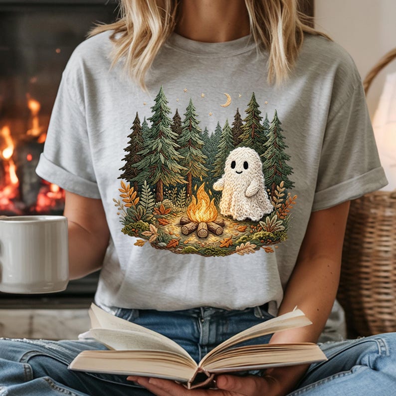 May include: A light gray t-shirt featuring a graphic of a friendly ghost sitting by a campfire in a forest. The design includes pine trees, a crescent moon, and autumn leaves. The ghost is white, and the flames are orange and yellow.