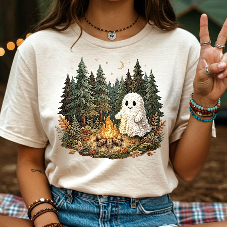 May include: A cream-colored t-shirt featuring a graphic of a friendly ghost sitting by a campfire in a forest. The illustration includes green pine trees, a crescent moon, and autumn leaves. The ghost is white and appears to be knitted.