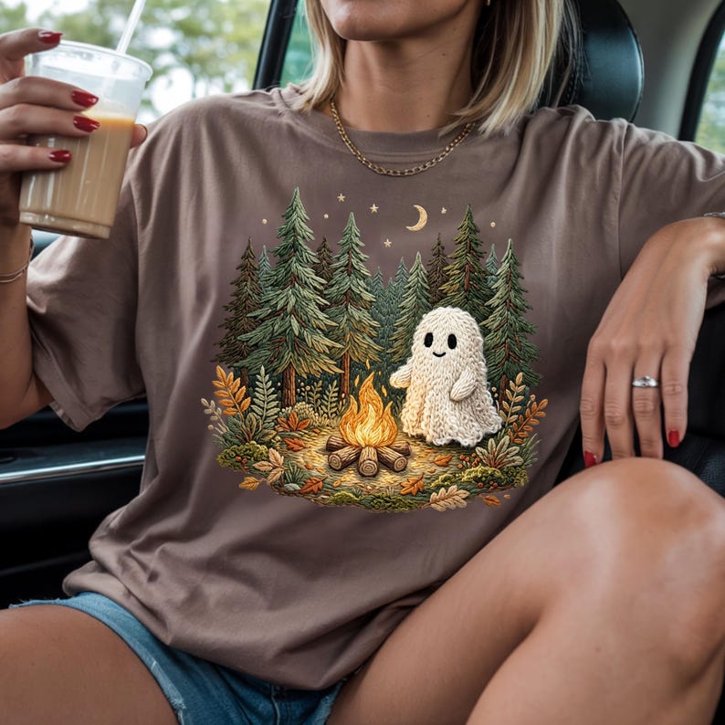 May include: A brown t-shirt featuring a whimsical illustration of a campfire scene with a friendly ghost, trees, and a crescent moon. The design includes a campfire with flames, a knitted ghost, and a forest backdrop. The shirt is a neutral color.