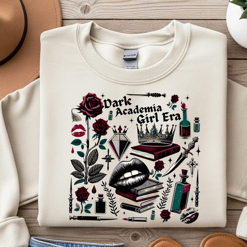 K&ouml;nnte beinhalten: Cremefarbenes Sweatshirt mit einem Grafikdesign. Das Design zeigt den Text "Dark Academia Girl Era" sowie Rosen, eine Krone, B&uuml;cher, einen Diamanten, Lippen und andere dekorative Elemente in Burgunderrot, Schwarz und T&uuml;rkis.