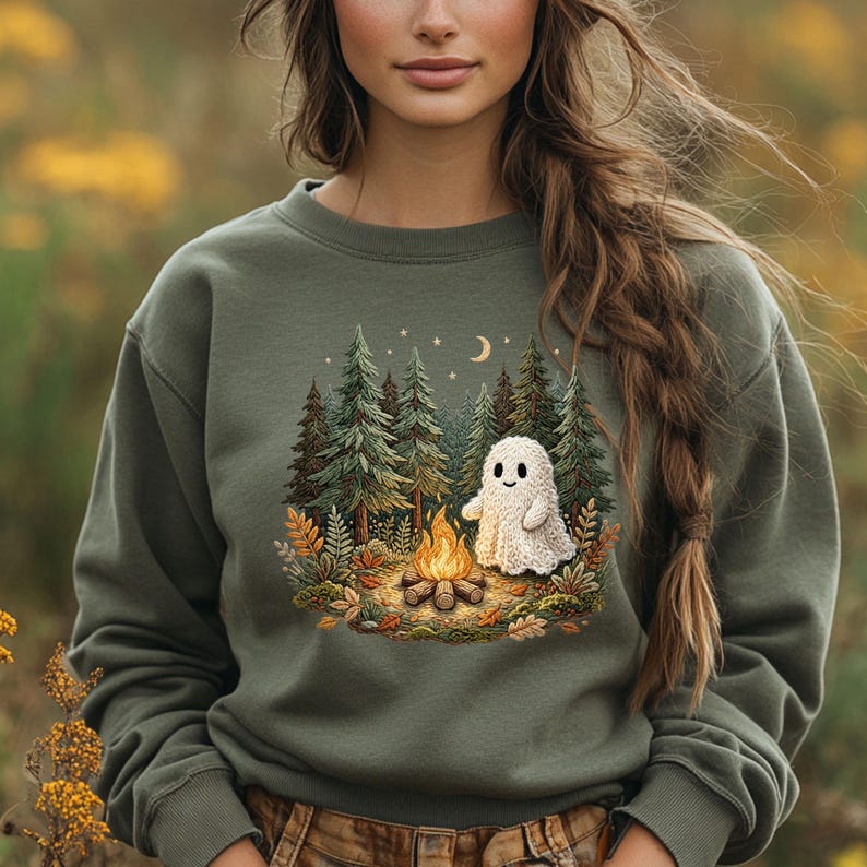 May include: Olive green sweatshirt featuring a whimsical illustration of a campfire scene with a friendly ghost, pine trees, and a crescent moon. The design is centered on the front of the sweatshirt, creating a cozy, nature-inspired aesthetic. The sweatshirt has a crew neck and long sleeves.