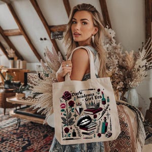 K&ouml;nnte beinhalten: Beige Canvas-Tragetasche mit einem grafischen Design mit dem Text "Dark Academia Girl Era". Das Design umfasst Rosen, einen Diamanten, eine Krone, B&uuml;cher und Lippen. Die Tasche hat zwei Griffe und wird von einer Person gehalten.