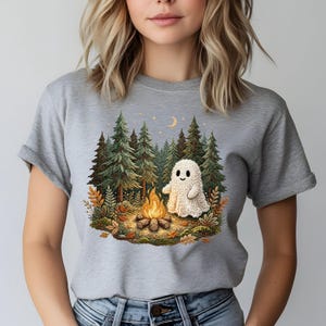 May include: A gray t-shirt featuring a whimsical illustration of a friendly ghost near a campfire in a forest setting. The design includes a crescent moon, stars, and autumn foliage. The ghost is made of a knitted material.