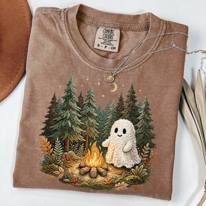 May include: A brown Comfort Colors t-shirt featuring a forest scene with a campfire and a friendly ghost. The graphic includes green trees, a crescent moon, and stars. A silver necklace with a pendant rests on the shirt.