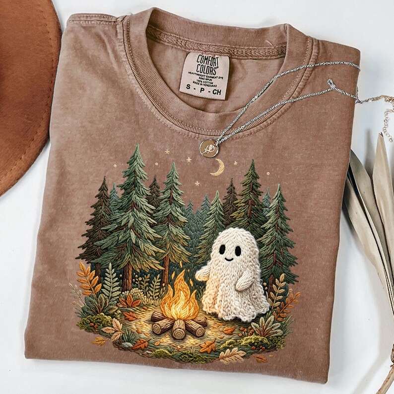 May include: A brown Comfort Colors t-shirt featuring a forest scene with a campfire and a friendly ghost. The graphic includes green trees, a crescent moon, and stars. A silver necklace with a pendant rests on the shirt.