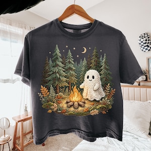 May include: Dark gray t-shirt featuring a whimsical illustration of a friendly ghost near a campfire in a forest setting. The design includes trees, a crescent moon, and stars, creating a cozy, outdoorsy theme.