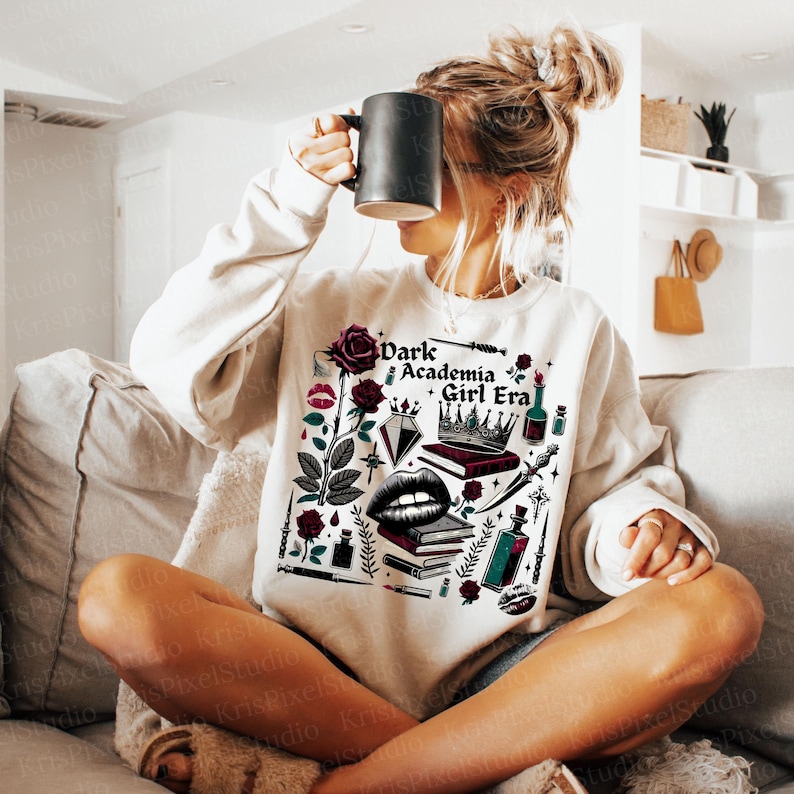 K&ouml;nnte beinhalten: Cremefarbenes Sweatshirt mit einem grafischen Design, das den Text "Dark Academia Girl Era" und Illustrationen von Rosen, B&uuml;chern, einer Krone und anderen gotisch inspirierten Elementen zeigt. Das Sweatshirt hat eine lockere Passform.