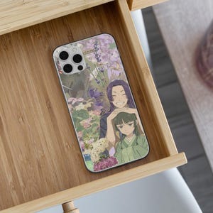 May include: A phone case with an anime-style illustration of two characters amidst flowers. The case has a light purple and green colour scheme, with a smiling character and another with a neutral expression. The case is inside a wooden drawer.