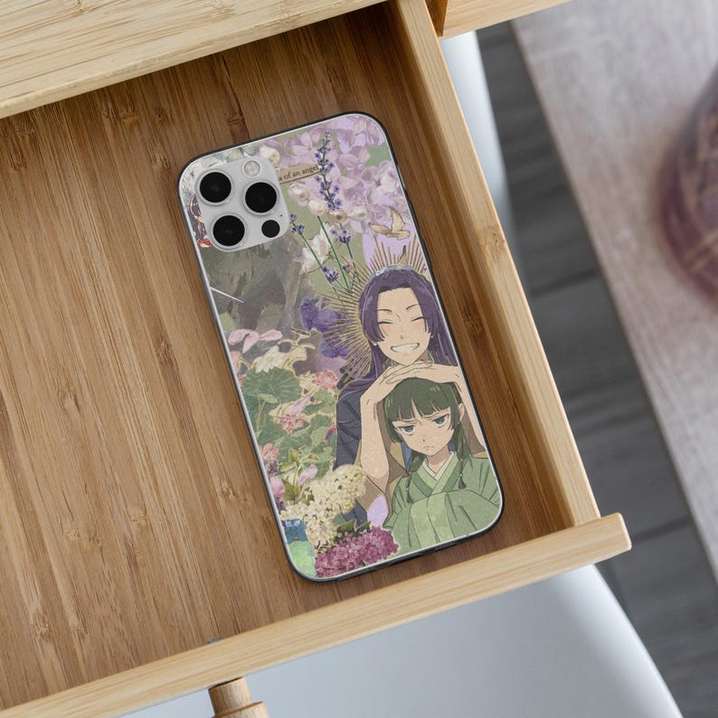 May include: A phone case with an anime-style illustration of two characters amidst flowers. The case has a light purple and green colour scheme, with a smiling character and another with a neutral expression. The case is inside a wooden drawer.