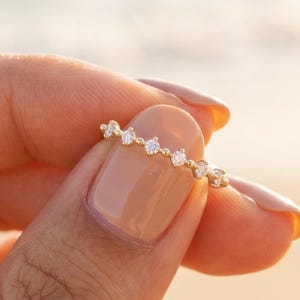 以下が含まれることがあります： 細いゴールドのリングで、バンドに沿って小さな丸い透明な石が繊細な列になって並んでいます。バンドは細いゴールドのワイヤーで作られており、石は均等な間隔で配置され、指に沿って繊細で連続した輝きのラインを作り出しています。