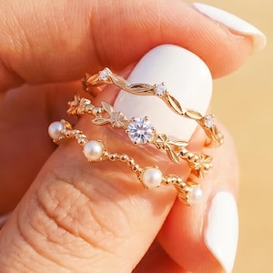 Könnte beinhalten: Drei goldene Ringe mit filigranen Designs sind auf einem Finger gestapelt. Der obere Ring hat kleine klare Steine, der mittlere Ring hat einen zentralen klaren Stein und florale Akzente, und der untere Ring hat kleine Perlen.