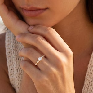 Puede incluir: Un anillo de oro con una gema grande, clara y redonda como pieza central. El anillo tiene una banda delicada con piedras de acento más pequeñas. El anillo está en el dedo de una persona, con una playa y el océano de fondo.