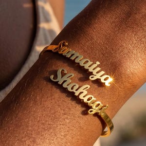 May include: A gold-toned metal cuff bracelet featuring two distinct lines of cursive script text. The top line displays the name "Sumaiya" and the bottom line displays the name "Shohagi" in a polished, reflective finish that catches the light.