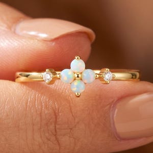 May include: A gold ring featuring a central cluster of four small, round, iridescent opals arranged in a cross shape, flanked by two tiny, clear, round-cut stones on either side of the band, set in a polished yellow gold metal setting.