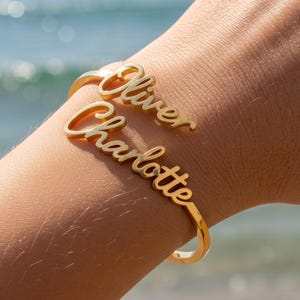 Pode incluir: Uma pulseira de aro aberto em tom dourado com dois nomes escritos em letra cursiva. O nome superior é Oliver e o nome inferior é Charlotte. O metal tem um acabamento polido e a pulseira foi concebida para envolver o pulso.