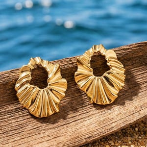 Peut inclure: Deux boucles d'oreilles dorées texturées avec un design à volants sont présentées sur un morceau de bois. Les boucles d'oreilles sont posées sur un fond de plage de sable et d'océan. Les boucles d'oreilles ont une forme circulaire.