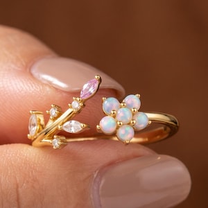 May include: A gold-toned open ring featuring a floral motif on one side with five round iridescent opal-like stones and a branch-like design on the other side, accented with small clear stones and a single marquise-cut pink stone.
