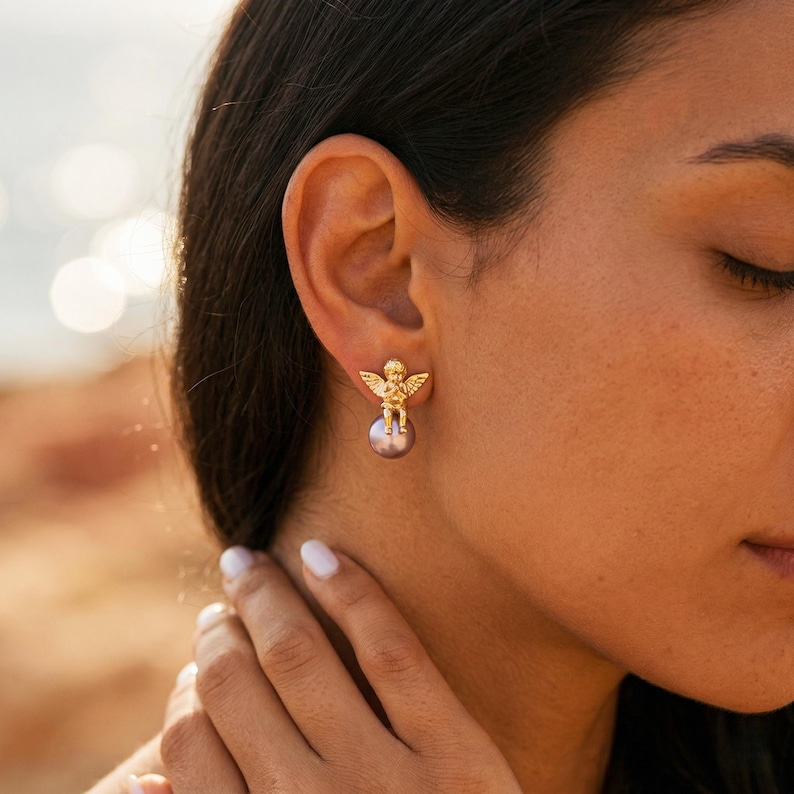 May include: Gold-colored angel earrings with a pearl. The earrings feature a small, detailed angel figure with outstretched wings atop a round, gray pearl. The earrings are shown on a person's ear.