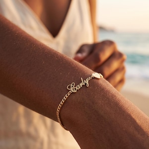 Könnte beinhalten: Goldfarbenes Armband mit dem Namen "Carly" in Schreibschrift. Das Armband hat einen kleinen Herzanhänger und ein Kettenglied-Design. Das Armband wird an einem Arm getragen, mit einer Strandszene im Hintergrund.