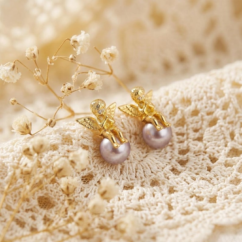 May include: A pair of gold-colored angel earrings with small wings and a small diamond on the head. The angels are perched on top of a light purple pearl. The earrings are displayed on a cream-colored lace fabric.