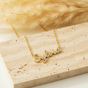 May include: A gold necklace featuring the name "Sylvia" in a cursive font, embellished with small, clear crystals. The necklace is presented on a light-toned, textured surface. Dried plants are visible in the background.