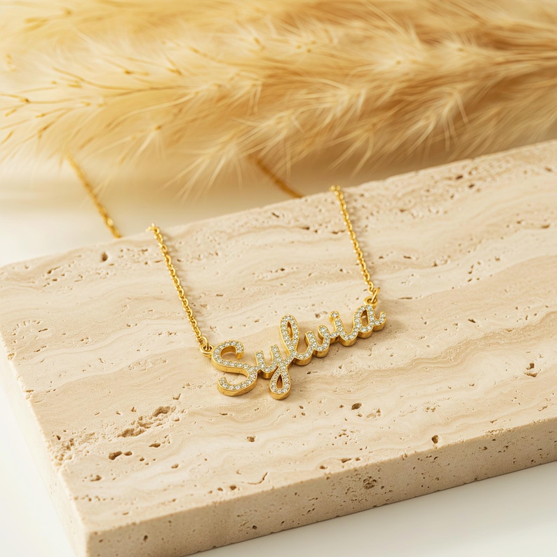 May include: A gold necklace featuring the name "Sylvia" in a cursive font, embellished with small, clear crystals. The necklace is presented on a light-toned, textured surface. Dried plants are visible in the background.